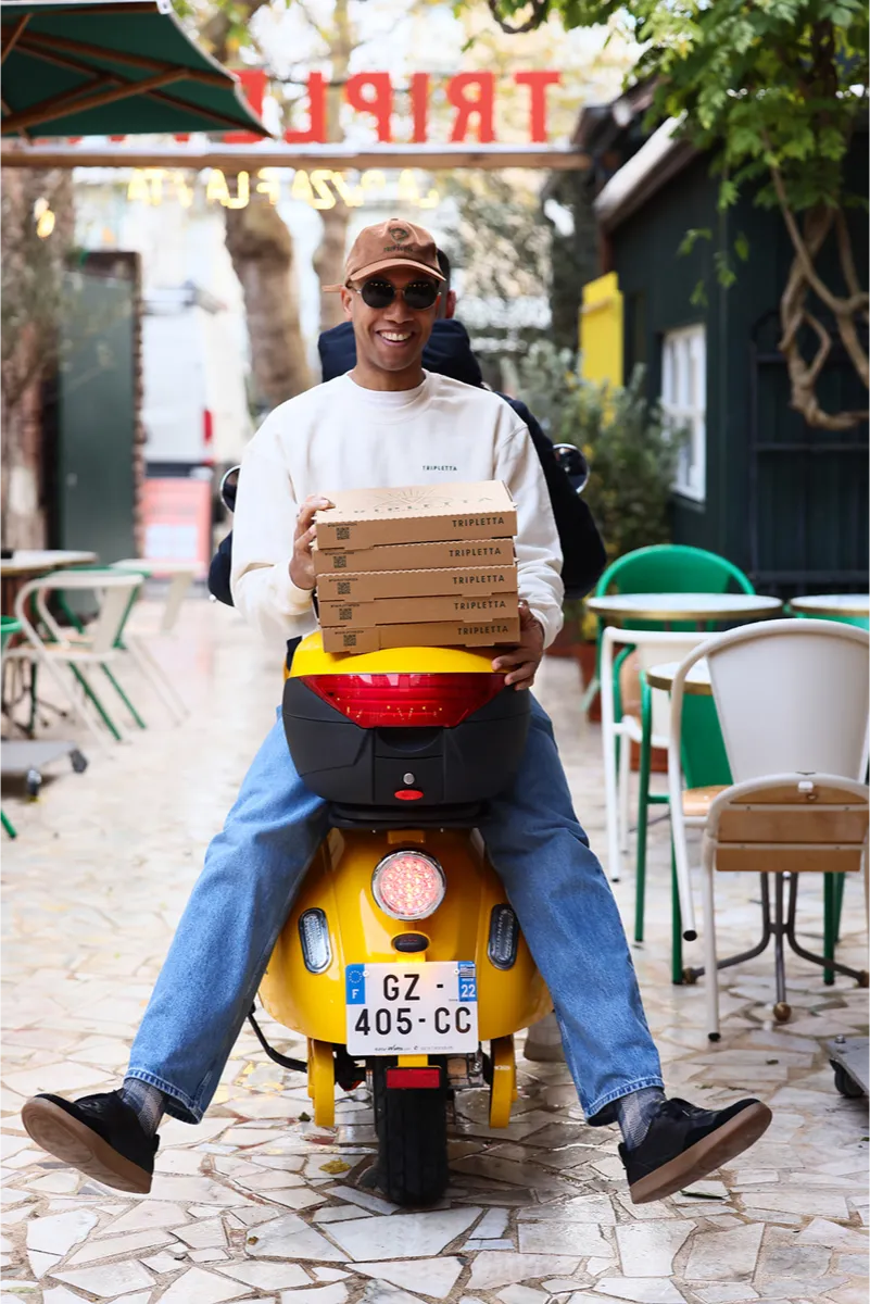 Livreur de pizza sur scooter portant une casquette et des lunettes, prêt pour une livraison Rue Forno.